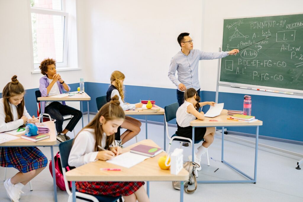 Students sitting at triangular desks in the classroom, there teacher is implementing the SIngapore approach to maths, explaining concepts on the blackboard