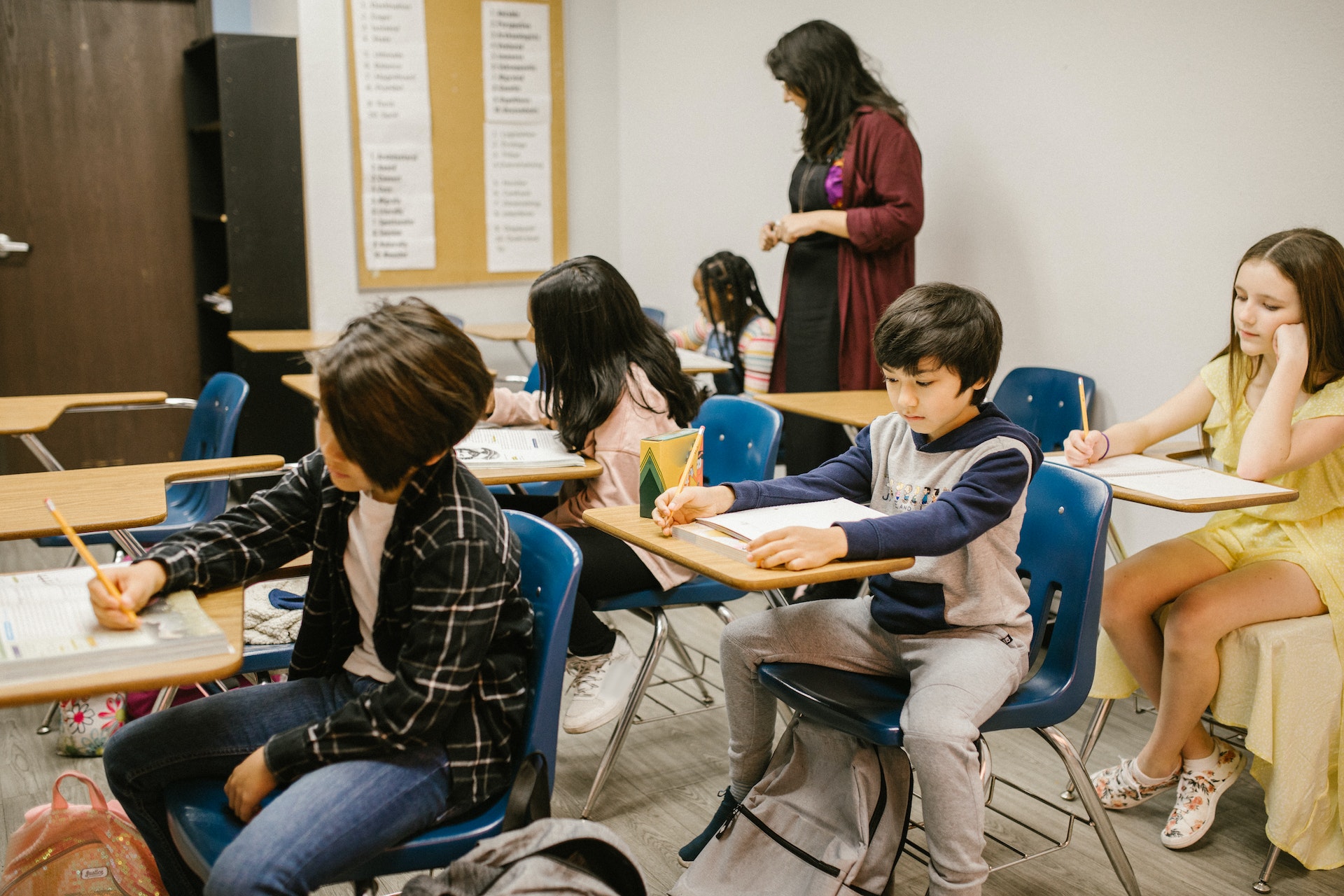 A group of learners working at their desks in the classroom, the teacher stands at the back of the class watching the students